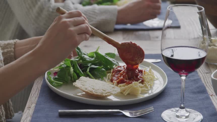A closeup of a dining table features a plate of pasta bread salad a glass of red wine and a hand adding sauce to the pasta with a spoon
