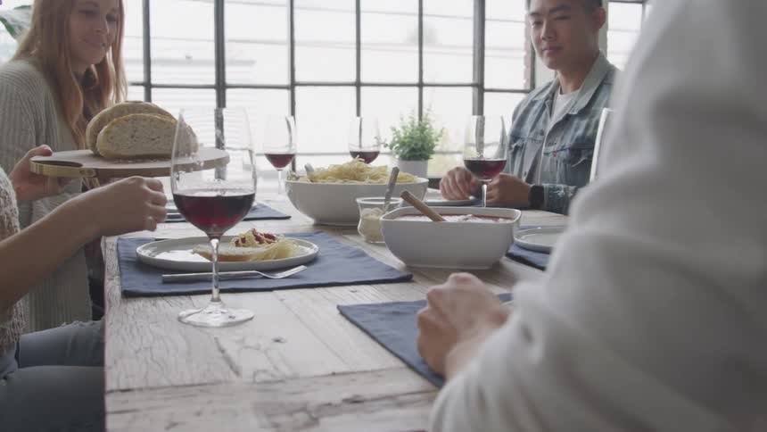 A group of friends sits around a wooden table set for a casual italian meal with pasta red wine and bread