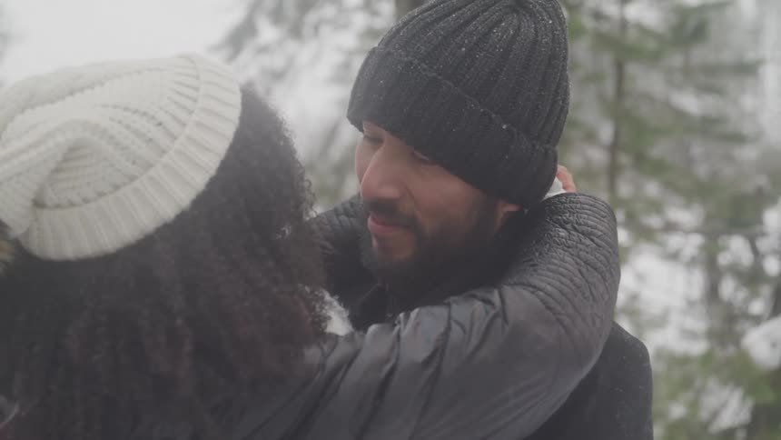Woman wraps her arms around the neck of a man while standing outdoors near the trees on a winter day