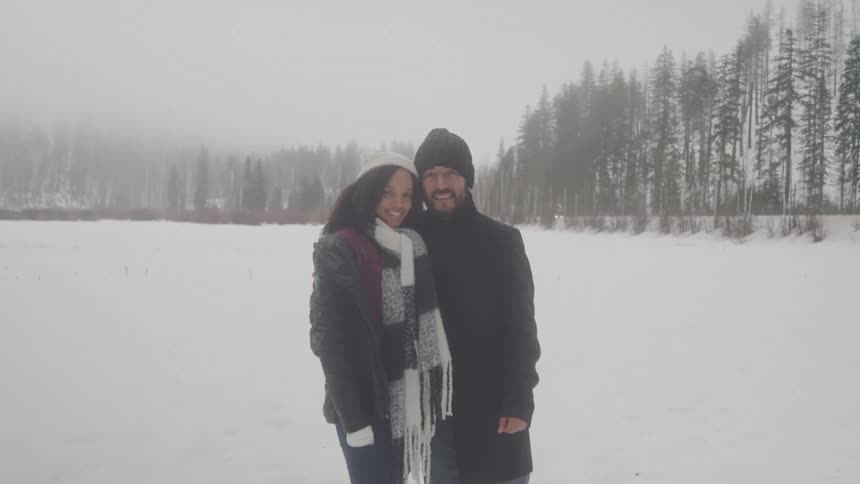 Couple wearing winter clothing standing together they have a side hug pose and both are smiling the scenery is outdoor in an open field that is surronded by trees and snow completely covers the ground
