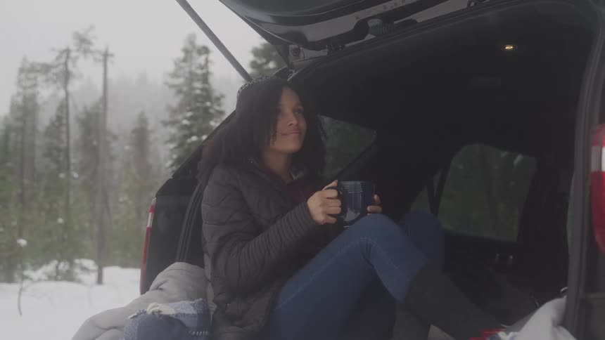 Woman sitting in back of suv in snow covered forest enjoying cup of coffee