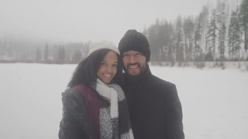 Couple poses smiling in the snow with an evergreen forest in the distance behind them