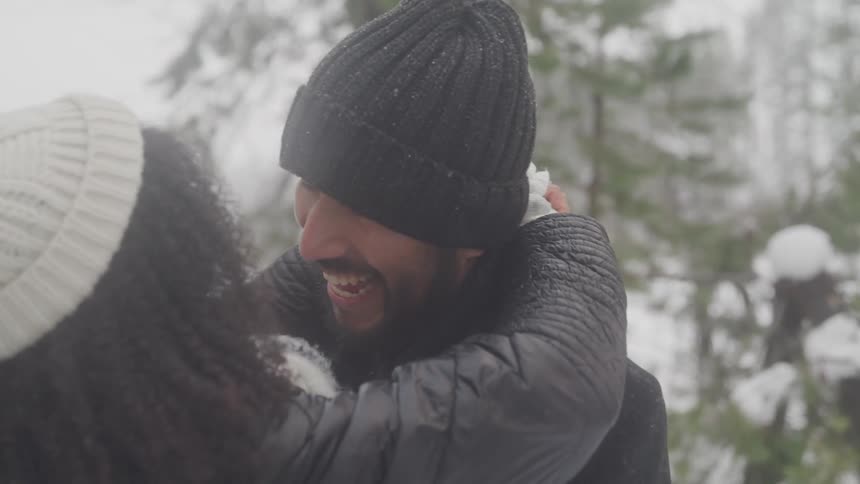 A happy couple embraces in winter clothes smiling and laughing while looking at each other in a snowy forest
