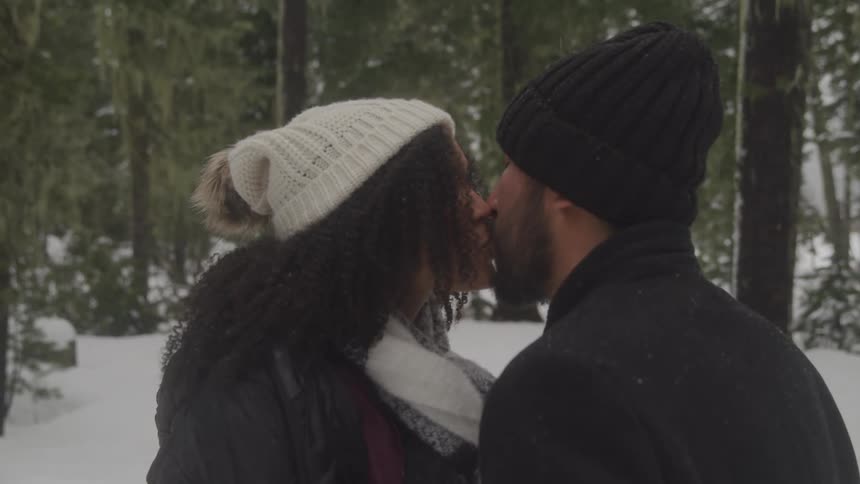 Couple bundled up in coats and beanies kissing in a snow covered forest