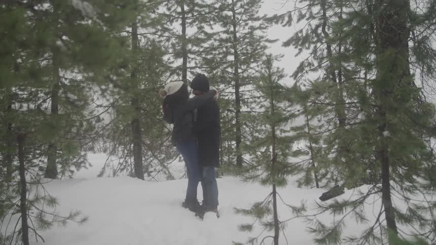 Man and woman bundled in winter clothing embrace amidst snow covered pine trees in a serene wintry landscape