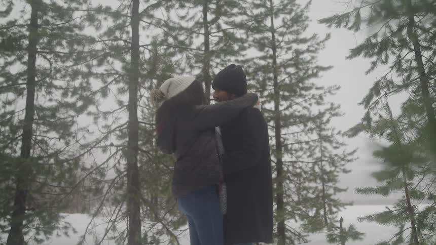 Man and woman dressed in winter clothing embrace amidst a snowy forest of tall pine trees