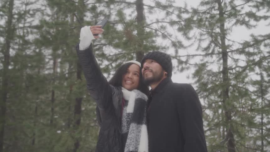 A couple takes a selfie in a snowy forest with the woman holding the phone up high