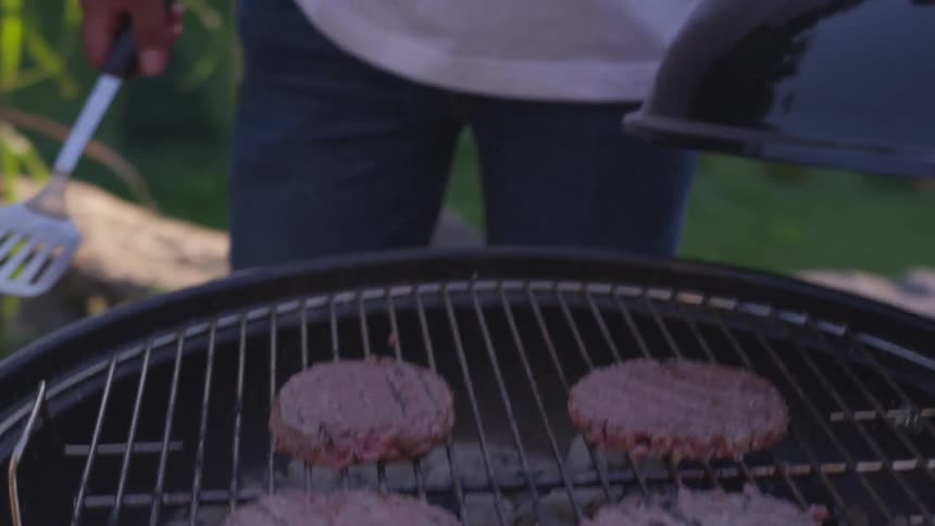 A man wearing a hat cooks hamburger patties on a charcoal grill at a barbecue