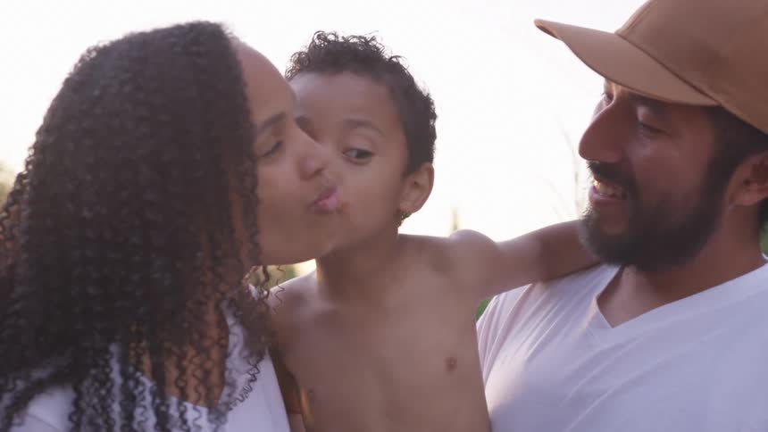 A couple holds a baby outdoors as the woman kisses the child and the father looks on with a smile under the warm light of either sunset or morning