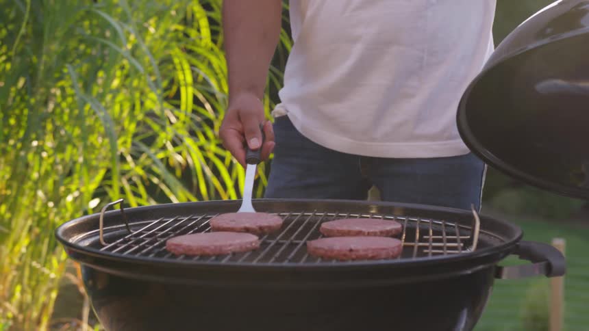 A man in a backyard holds a grill lid while using a spatula to flip burger patties cooking on a black barbecue pit