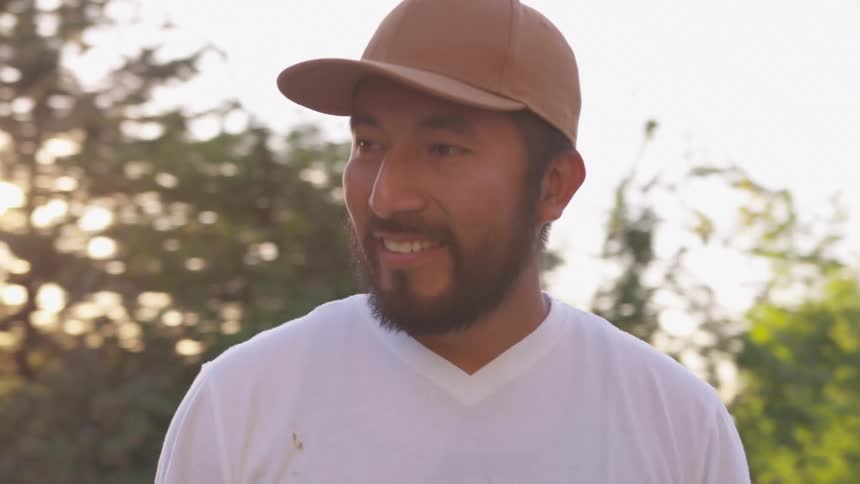An asian man with a beard in a brown baseball cap and white shirt looks up and to his right widely smiling at someone off camera with sunlight streaming through trees behind him