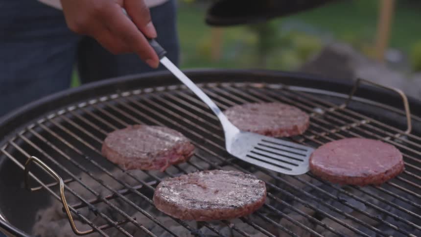 Holding a black grill lid a person uses a spatula to flip hamburger patties cooking on a charcoal grill in a backyard setting