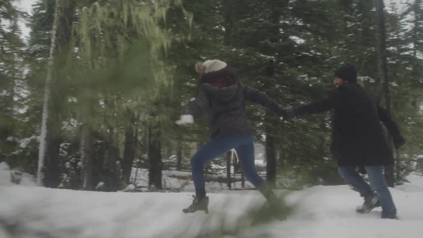 A man and a woman holding hands run through the snow in a forest