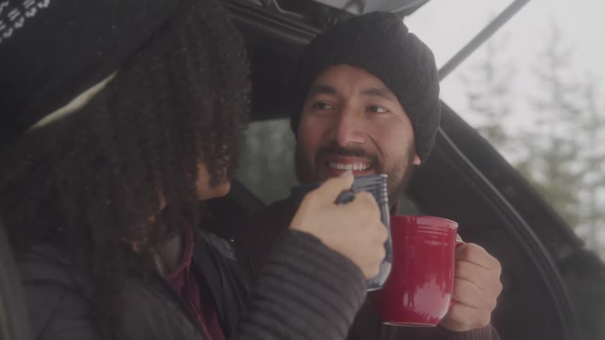 Male and female sitting in the back of a vehicle both are toasting eachother with mugs both people are smiling and seem to enjoy the moment the couple is outside and there are trees in the back ground and snow falling from the sky