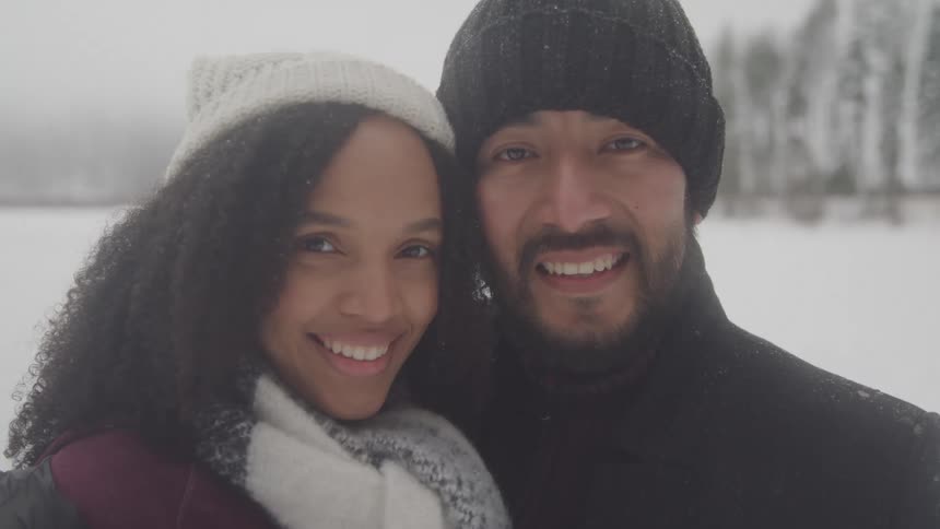 A smiling couple bundled up in winter hats and scarves poses for a snowy outdoor portrait