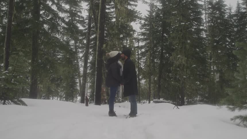 A man and woman slowly lean in to kiss standing in a snowy area with trees