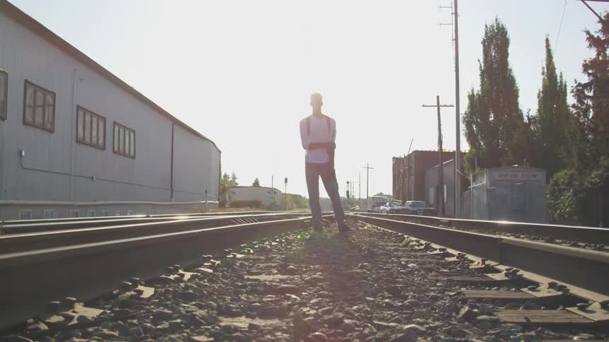 A silhouetted figure stands on railroad tracks stretching into the distance under a bright sunlit sky