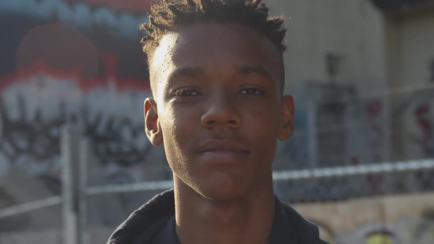 A young man with short textured hair is captured with a subtle smile looking directly at the camera against a blurred urban backdrop