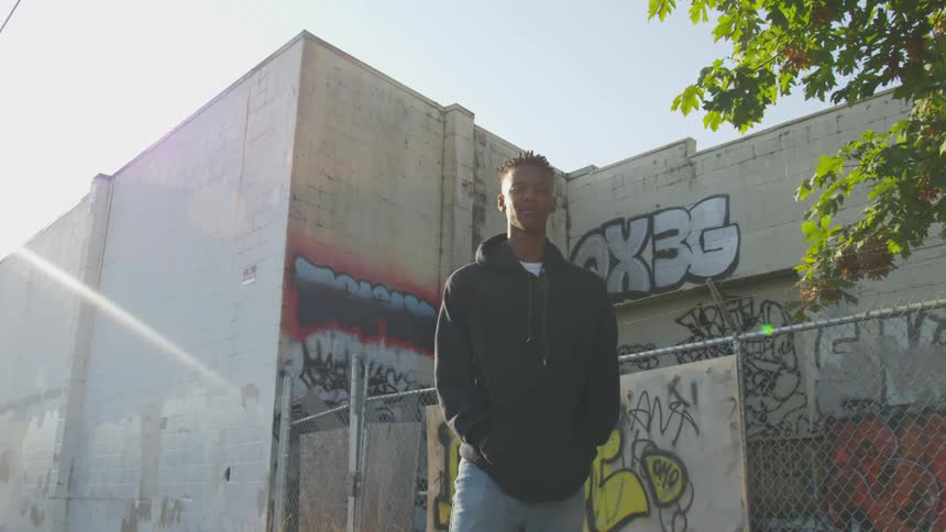 A young black man in a hoodie stands in front of a building covered in graffiti framed against a bright sunny sky