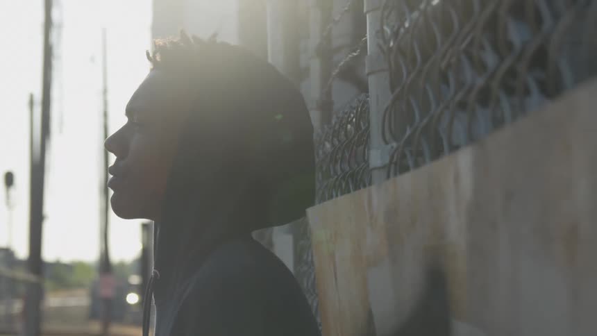 A teenager wearing a hoodie reclines against a wired fence with sunlight streaming over a cityscape in the background