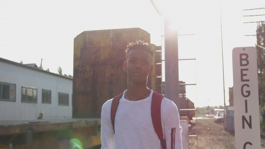 The black man is standing in front of an electric pole that is beside a train that is moving