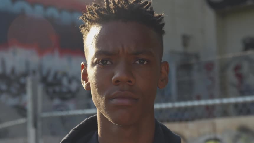 A young person with short textured hair looks directly at the camera with a serious expression