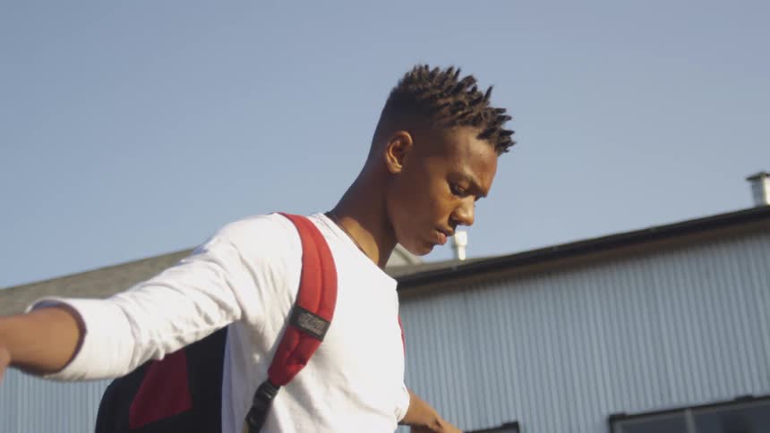 A teen wearing a white longsleeve shirt and a red backpack walks carefully along an outdoor path beside a corrugated metal structure under a vivid blue sky