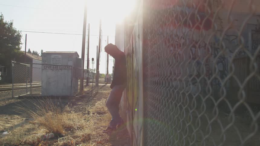 A person stands hunched and leaning against a wall near a chain link fence in a sunlit industrial looking area