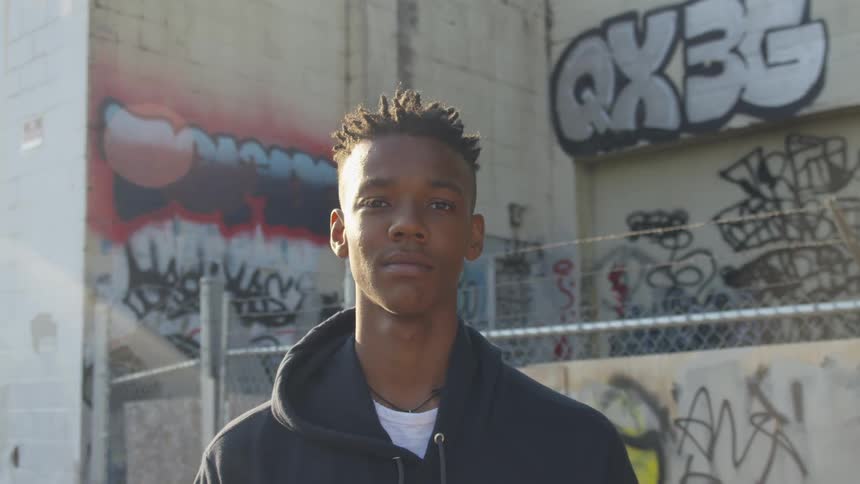 A young man in a black hoodie stands against a graffiti covered wall
