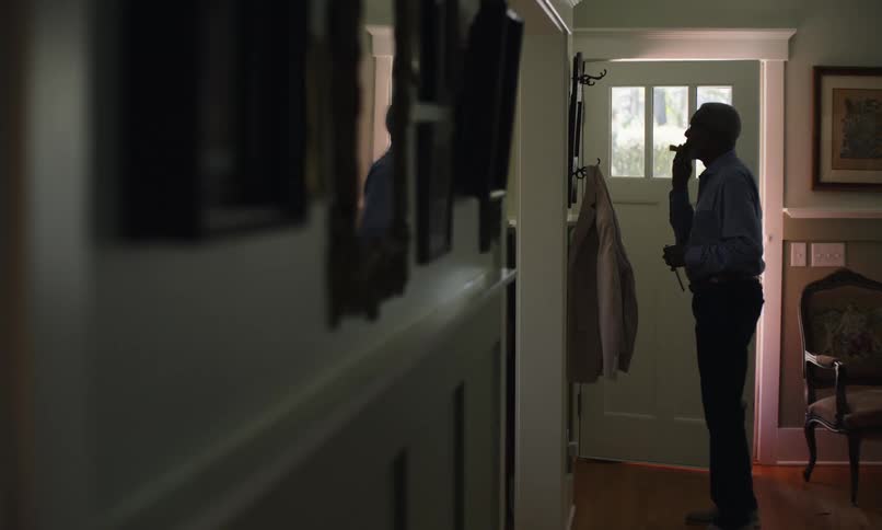 An elderly man stands in a hallway near his front door facing a mirror and carefully combing both his hair and beard while looking at his reflection