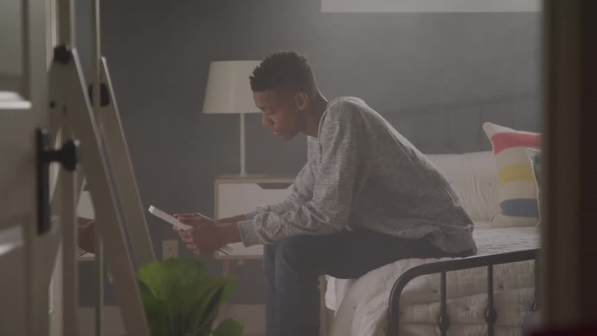 A young man with short dark hair sits on the edge of a bed looking down at a tablet in a dimly lit room