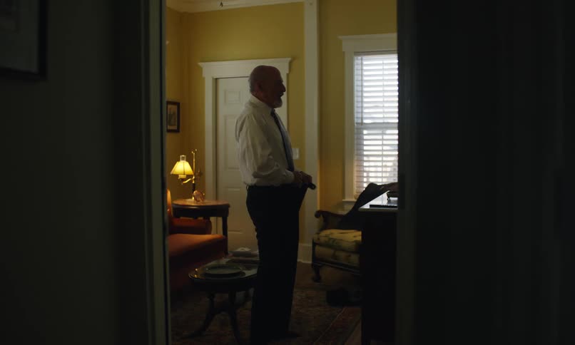 Older man in a dim lityellow painted room room tucking in his button up shirt into his pants he adjusts his tie and shirt when he is finsihed tucking his shirt in once he is done he takes a final look at himself