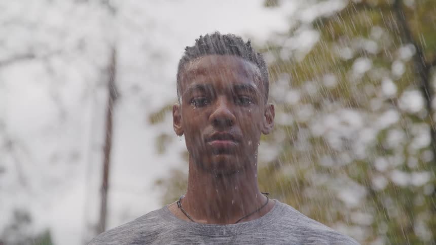 A young man with short dark hair stands facing the camera with rain falling around him creating a blurred textured effect against a backdrop of trees