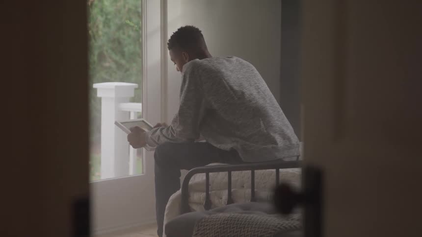 A man with a gray sweater and dark pants sits on a bed facing a window and using a tablet device