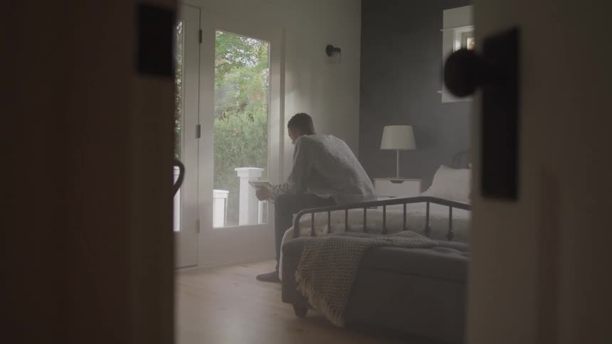 Man sits on the edge of a bed and looks at an electronic device