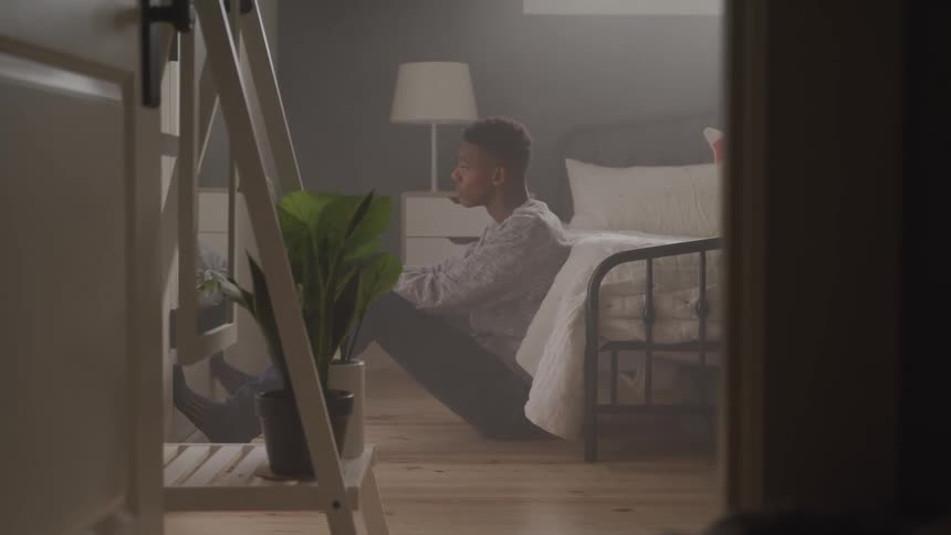 Boy sitting on the floor leaning against a bed very distressed