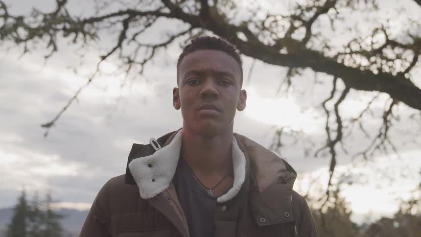 A young man with short dark hair stares directly at the camera with a serious expression against a backdrop of bare tree branches and a cloudy sky