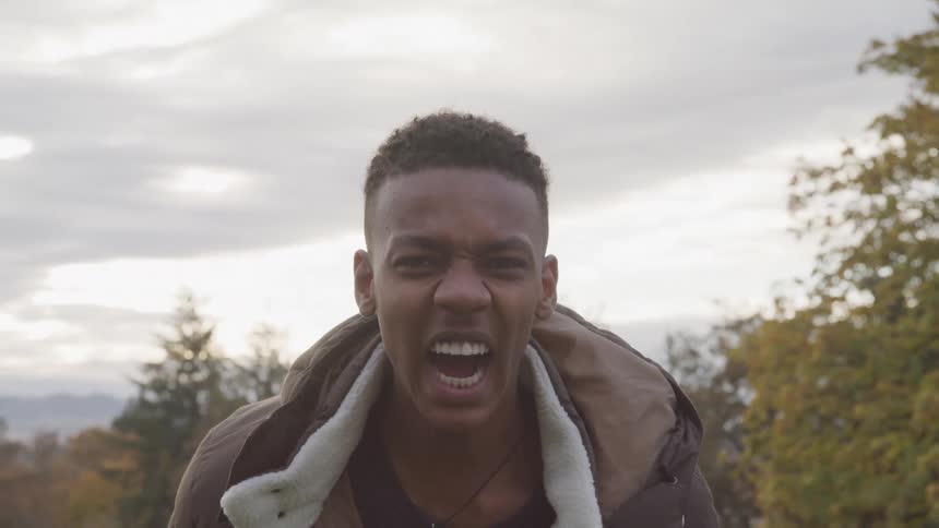 A young man with short dark hair wearing a brown jacket shows him shouting with an intense emotional expression against a blurred background of trees and an overcast sky
