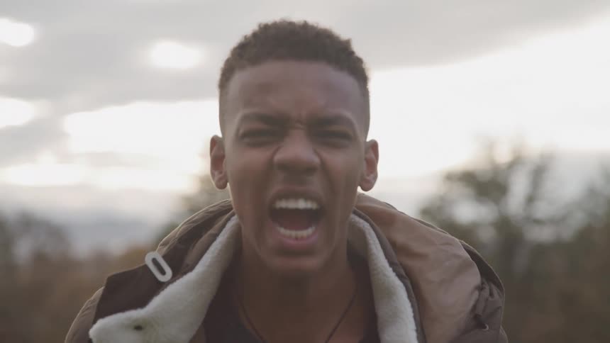 A young man with short dark hair wearing a brown jacket shows him shouting with an intense expression against a cloudy sky backdrop