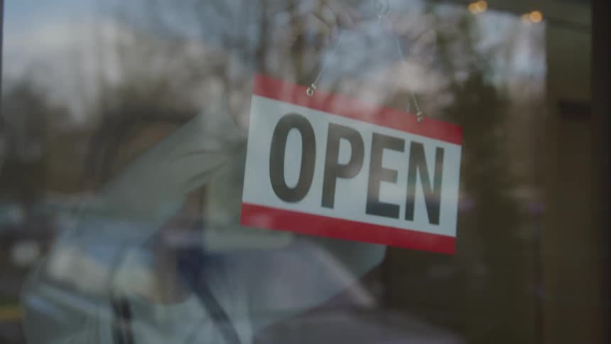 A woman inside a store turns the open sign around as she smiles