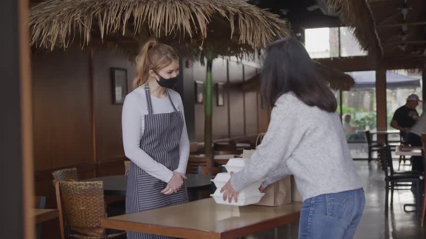 A woman picks up a stack of white takeout containers from a counter in a tiki themed restaurant leaving brown paper bags behind