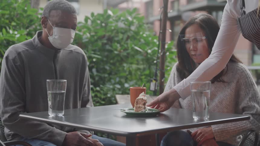 Couple wearing face covers sitting in patio outdoors being served a slice of cake by the waitress