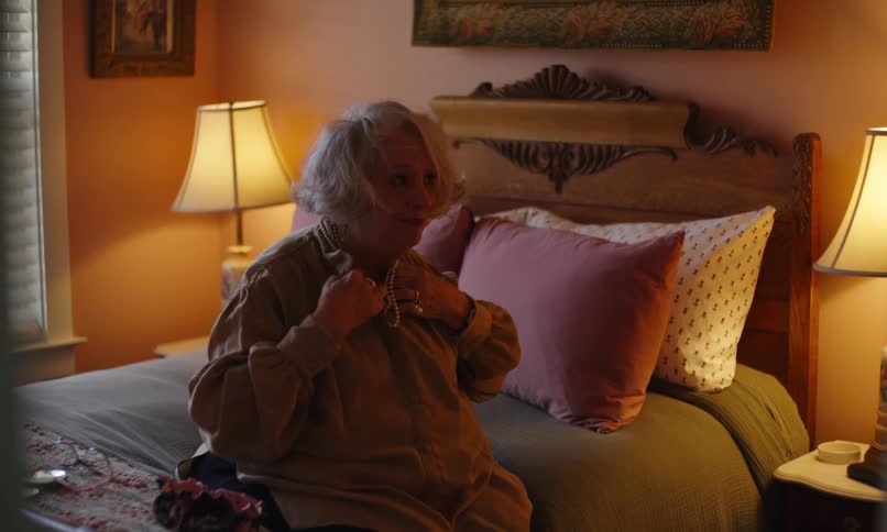Elderly woman puts on a necklace while sitting on her bed