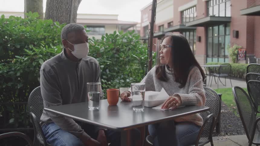 2 people with masks sitting at a table in a bar talking
