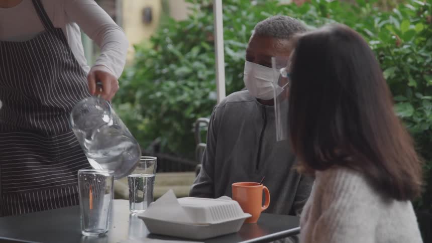 A waiter in a blue and white apron serves glasses of water to a couple dining outside