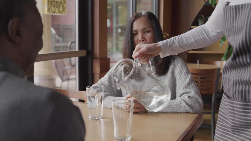 The man and woman are sitting at the table while the waitress is pouring their water into the tall glasses that are on the table in front of them
