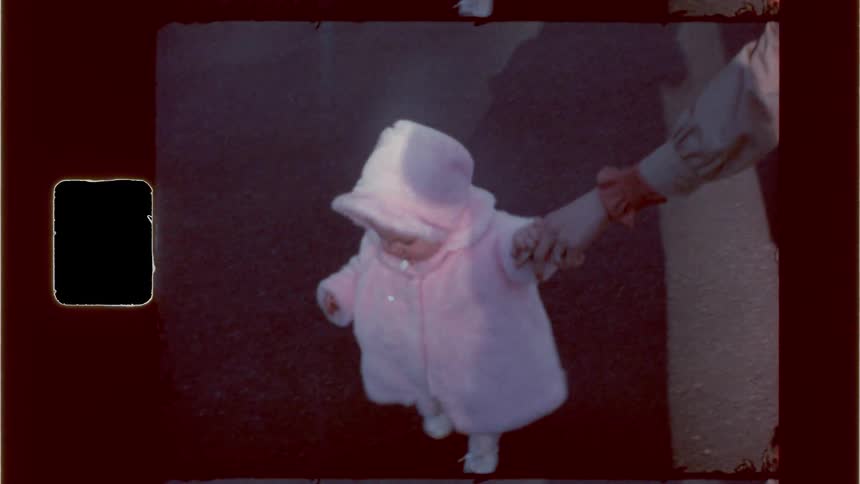 Baby in a pink furry coat and bonnet toddling about outdoors with mothers help