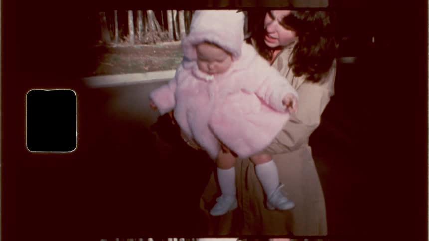 A baby in a pink hooded coat holding hands with the mother against a dark grainy background with silhouetted figures