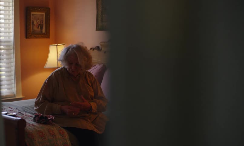 An elderly woman sits on her bed in a warm toned room her figure partially obscured by the foreground
