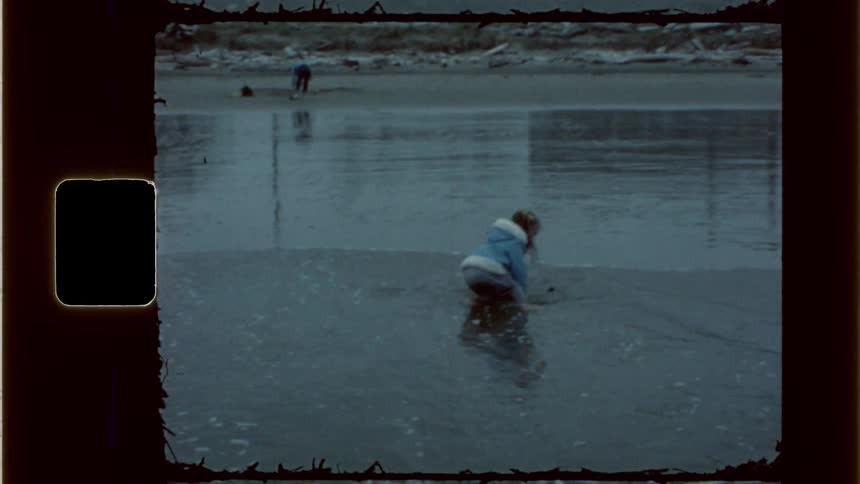 The little girl is running with the waves and falls down but she jumjps right back up and continues to head towards her parent that is standing on the beach